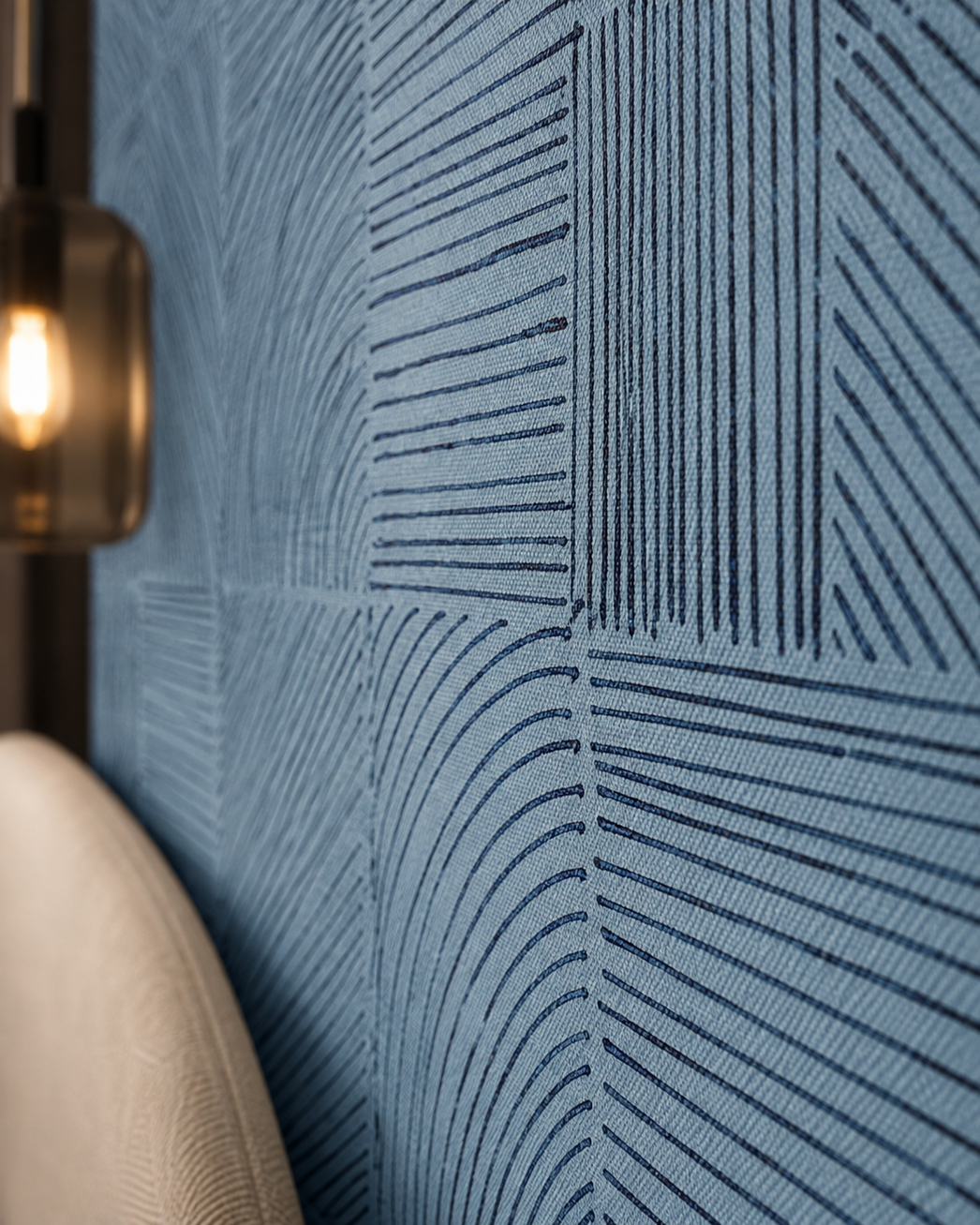 A close-up of textured blue wallpaper with a geometric pattern of intersecting lines in varying directions, with a warm glowing wall lamp visible on the left and a beige upholstered chair edge at the bottom left.